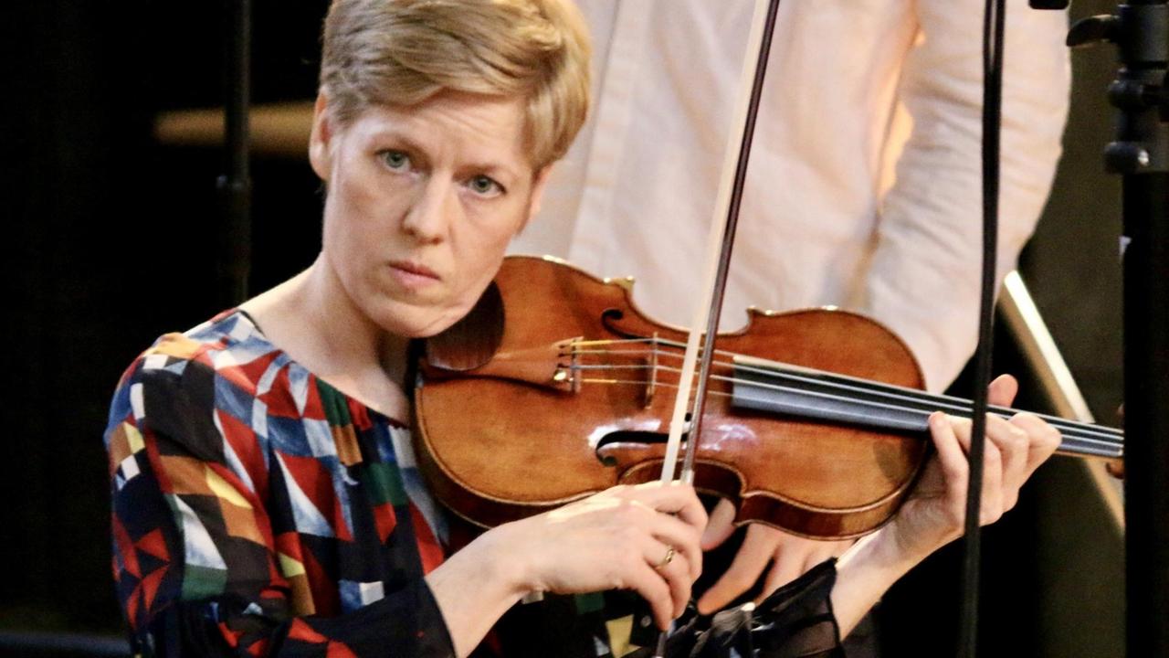 Eine blonde Frau mit kurzen Haare, buntem Oberteil und fokussiertem Blick spielt Geige. Eine blonde Frau mit kurzen Haare, buntem Oberteil und fokussiertem Blick spielt Geige.