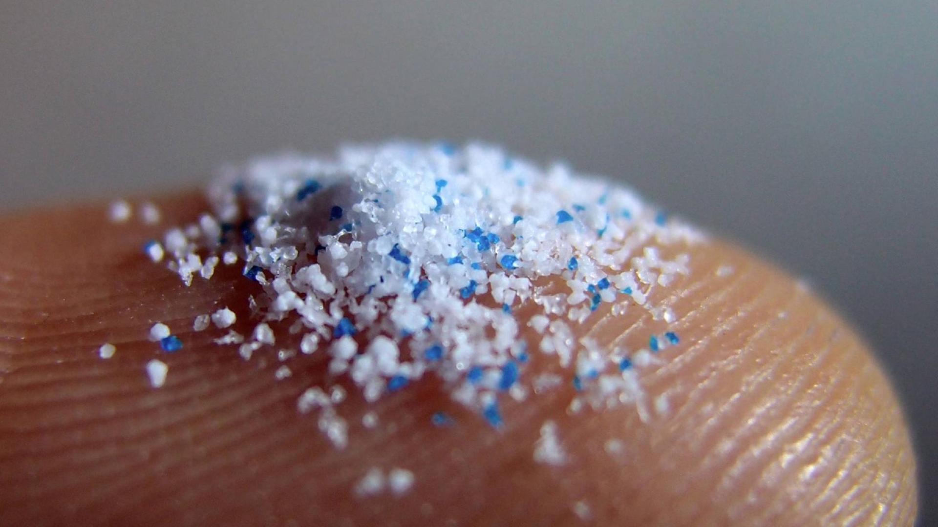 Blaue und weiße Mikroplastik-Partikel auf einer Fingerspitze.