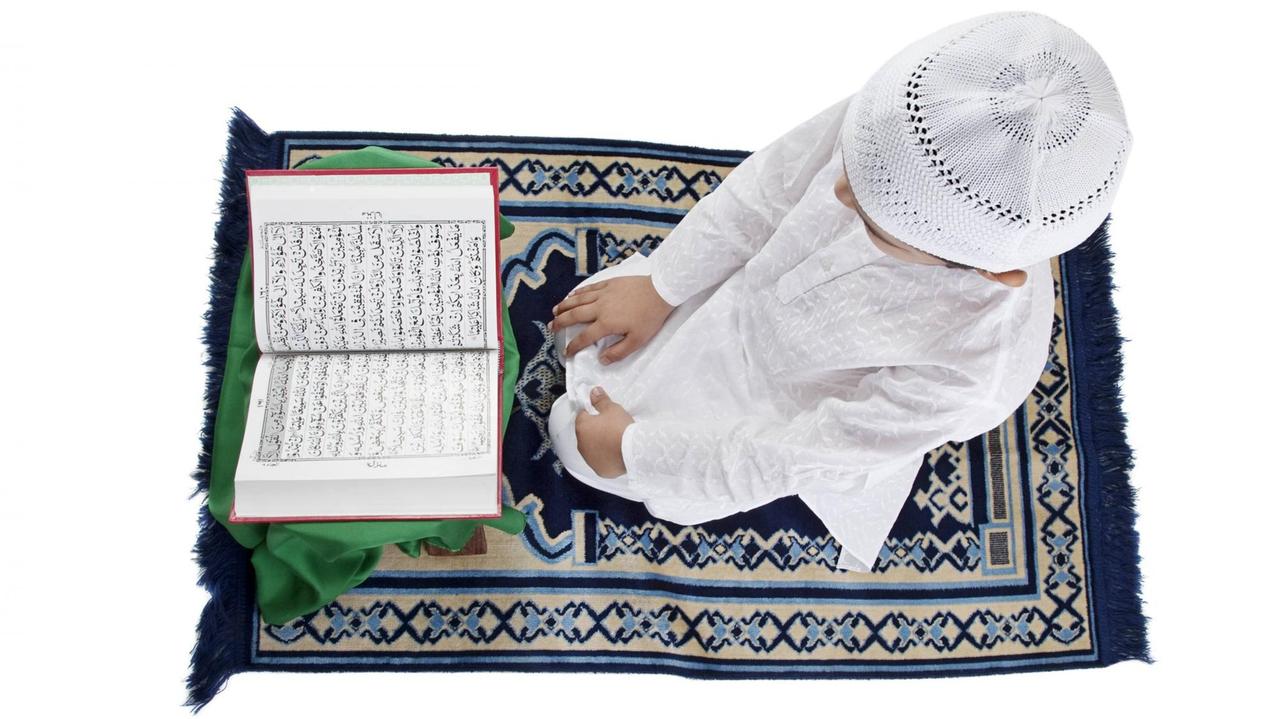 Ein muslimischer Junge in weißem Gewand und mit Häkelkappe sitzt auf einem Gebetsteppich und liest den Koran. Ein muslimischer Junge in weißem Gewand und mit Häkelkappe sitzt auf einem Gebetsteppich und liest den Koran.