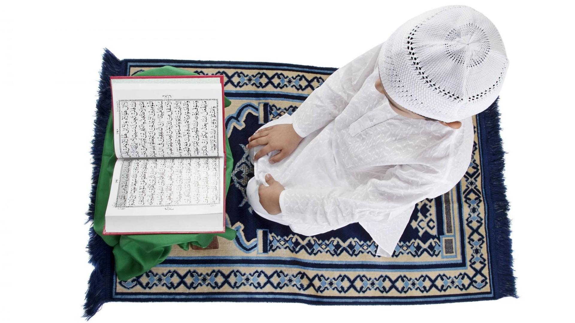 Ein muslimischer Junge in weißem Gewand und mit Häkelkappe sitzt auf einem Gebetsteppich und liest den Koran. Ein muslimischer Junge in weißem Gewand und mit Häkelkappe sitzt auf einem Gebetsteppich und liest den Koran.
