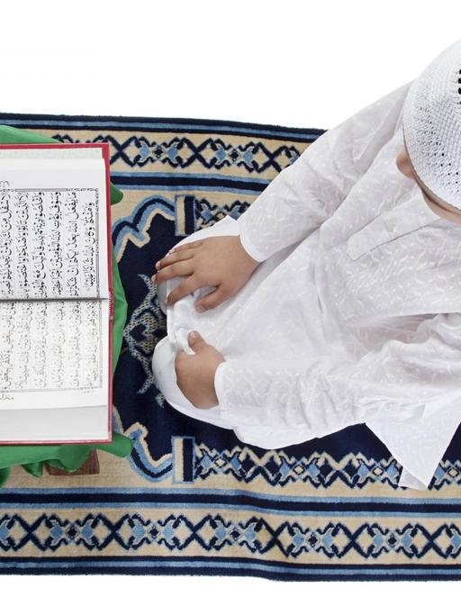 Ein muslimischer Junge in weißem Gewand und mit Häkelkappe sitzt auf einem Gebetsteppich und liest den Koran.