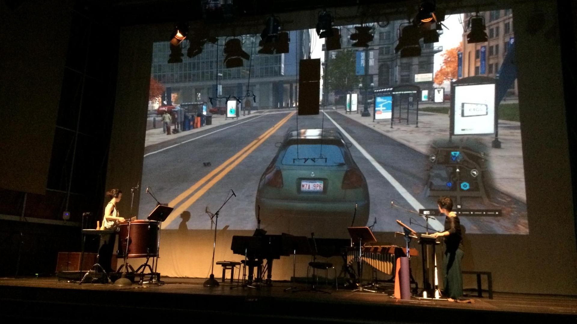  Zwei Musikerinnen stehen auf einer Bühne, im Hintergrund eine große Leinwand, auf der ein animiertes Autovideo zu sehen ist.