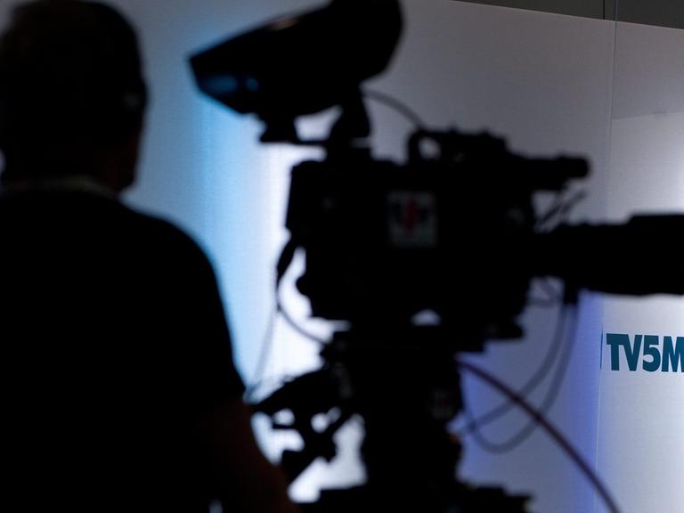 Schattenriss einer Fernsehkamera und ihres Kameramanns vor einer weißen Wand mit dem Logo von TV5 Monde.