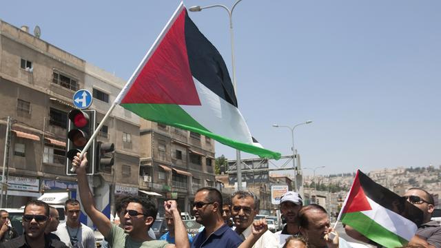 Menschen auf einer Straße vor Häusern, halten die palästinensische Flagge hoch, Arme in der Luft, rufen.