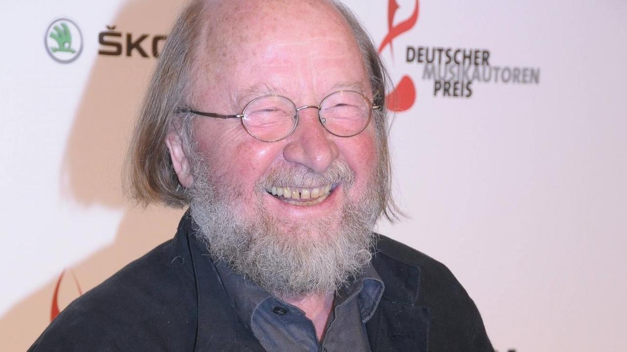 Ein Mann steht bei einer Preisverleihung auf dem roten Teppicht vor einer Präsentationswand und lacht herzlich Richtung Fotografen. Ein Mann steht bei einer Preisverleihung auf dem roten Teppicht vor einer Präsentationswand und lacht herzlich Richtung Fotografen.
