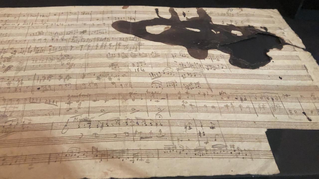 Ein dicht beschriebenes Notenblatt zeigt ein Loch, das mit einem großflächigem Tintenfleck umgeben ist. Ein dicht beschriebenes Notenblatt zeigt ein Loch, das mit einem großflächigem Tintenfleck umgeben ist.
