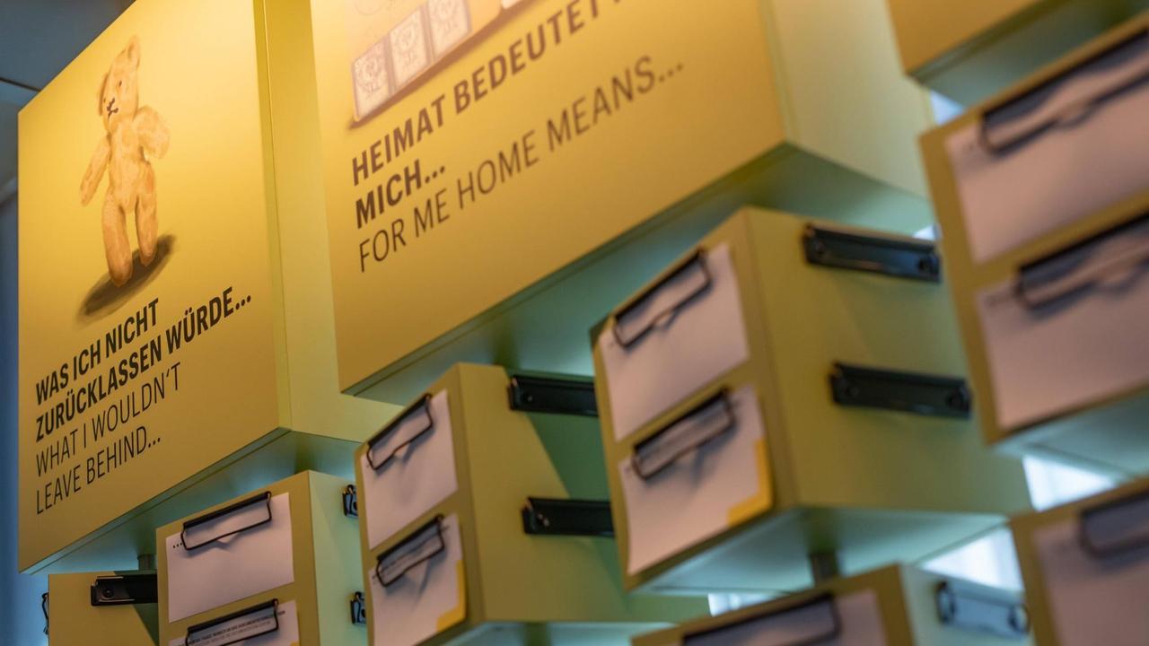 Einblick in die Ausstellung des Dokumentationszentrums Flucht, Vertreibung, Versöhnung im Deutschlandhaus in Berlin. Auf gelben quadratischen Flächen stehen die unfertigen Sätze: "Heimant bedeutet für mich..." und "Was ich nicht zurücklassen würde...". Darunter sind leere Zettel an gelben drehbaren Kästen angebracht. 