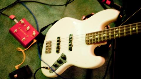 Ein weißer E-Bass (Model Fender Jazz Bass) steht neben einem Effektgerät auf einem Teppich in einem Proberaum.