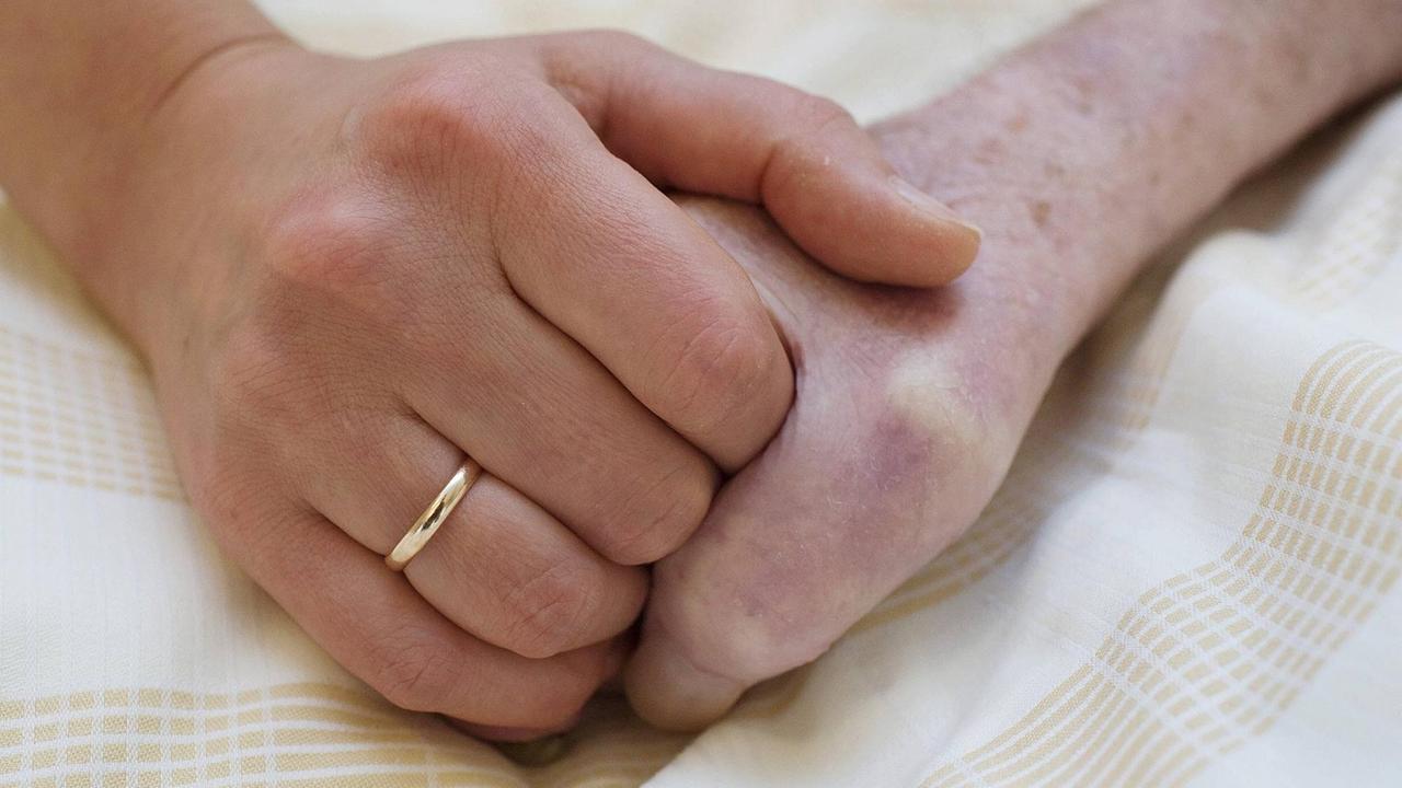 Zwei Hände halten sich umschlossen auf einem Laken. Die Linke Hand trägt einen Ehering. Zwei Hände halten sich umschlossen auf einem Laken. Die Linke Hand trägt einen Ehering.