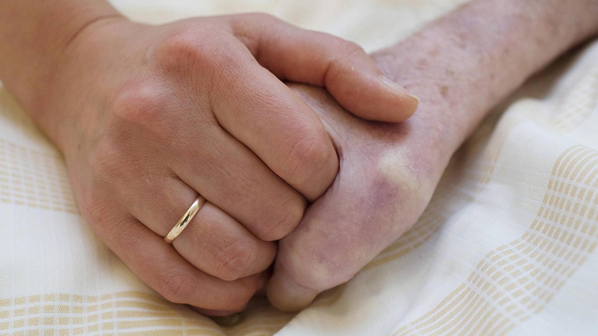 Zwei Hände halten sich umschlossen auf einem Laken. Die Linke Hand trägt einen Ehering. Zwei Hände halten sich umschlossen auf einem Laken. Die Linke Hand trägt einen Ehering.