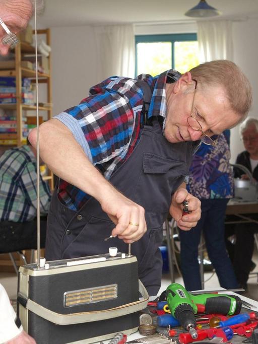 Ein Mann repariert ein altes Radio bei einem Repair Café Treffen. Repair Cafés sind ehrenamtliche Treffen, bei denen die Teilnehmer alleine oder gemeinsam mit anderen ihre kaputten Dinge reparieren.