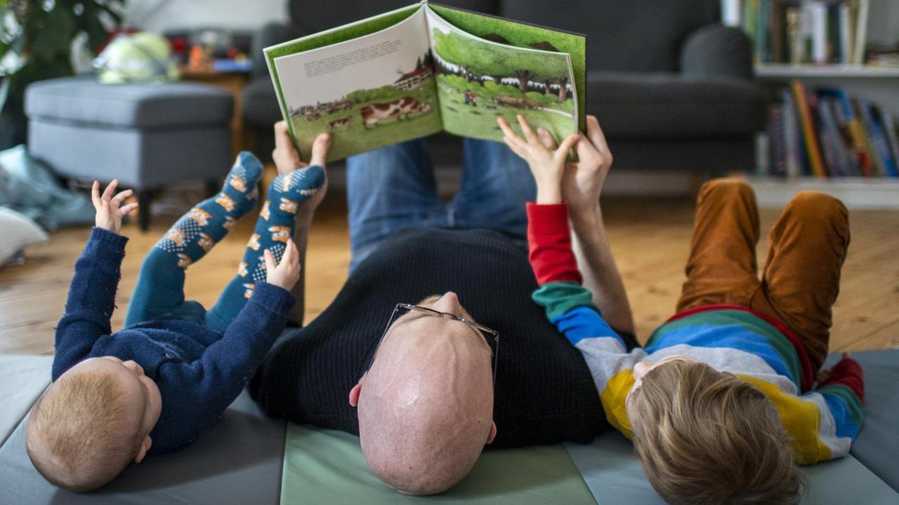 Ein Vater liest mit seinen beiden Kindern auf dem Boden liegend ein Buch. Ein Vater liest mit seinen beiden Kindern auf dem Boden liegend ein Buch.