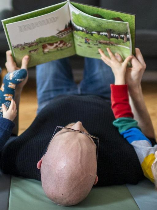 Ein Vater liest mit seinen beiden Kindern auf dem Boden liegend ein Buch.