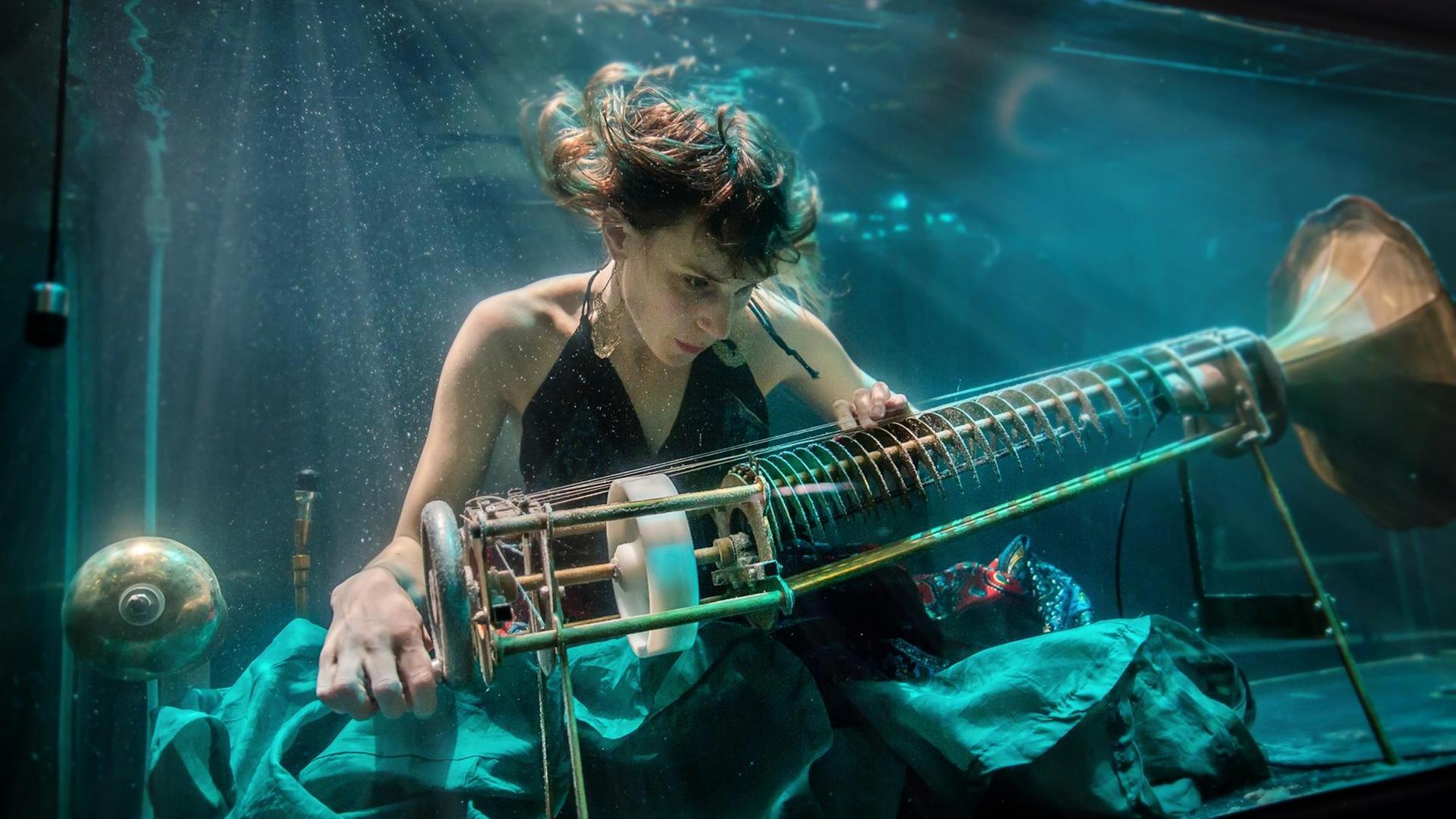 Die Künstlerin Nana Bech ist in einem Wasserbecken zu sehen. Sie spielt ein speziell konstruiertes Instrument namens Rotacorda. Links hat eine Kurbel, rechts einen Trichter wie vom Grammophon bekannt.