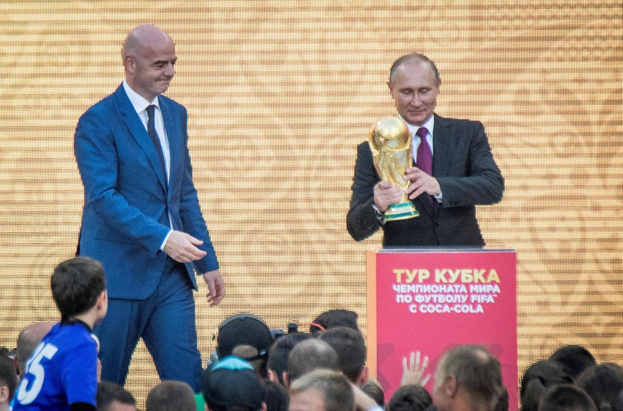 Putin (re.) hält eine goldene Trophäe, Infantino kommt von links auf ihn zu. Vor der Bühne stehen Zuschauer. Putin (re.) hält eine goldene Trophäe, Infantino kommt von links auf ihn zu. Vor der Bühne stehen Zuschauer.
