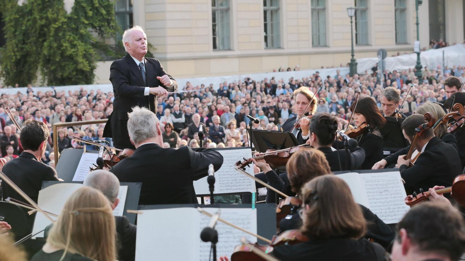 Ein Mann mit grauen Haaren steht inmitten eines Orchesters auf einem Pult und dirigiert. Ein Mann mit grauen Haaren steht inmitten eines Orchesters auf einem Pult und dirigiert.