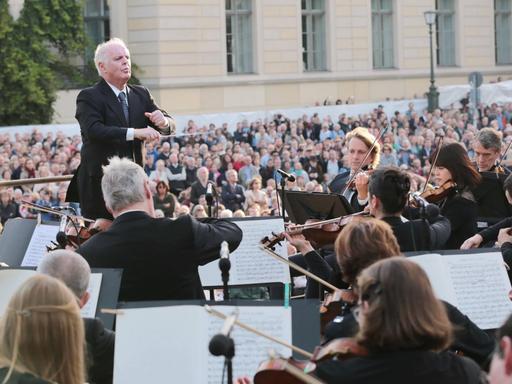 Ein Mann mit grauen Haaren steht inmitten eines Orchesters auf einem Pult und dirigiert.