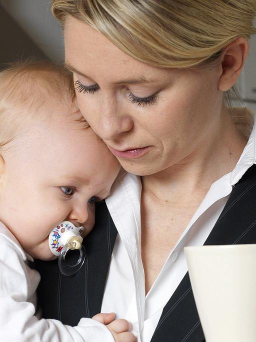 Eine Mutter im Business-Anzug mit ihrem Baby auf dem Arm, in der Hand eine Kaffeetasse.