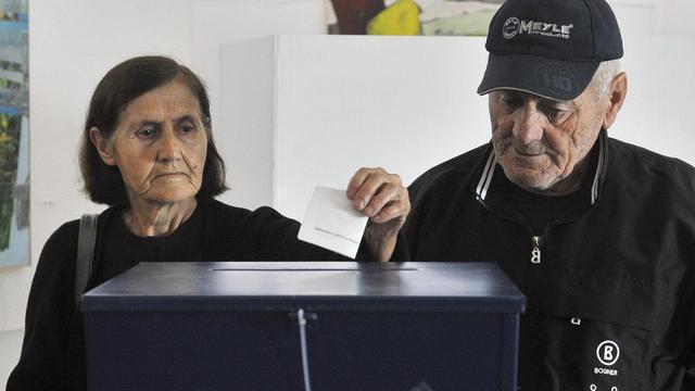 Ein älteres Ehepaar steht vor einer Wahlurne. Die Frau wirft einen Zettel hinein.
