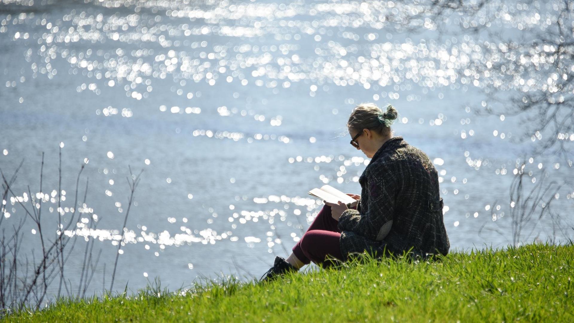 Eine Frau sitzt in Kassel am Ufer des Flusses Fulda und liest ein Buch.