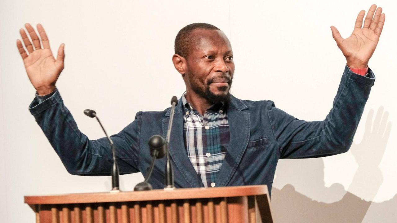 Olu Oguibe steht an einem Rednerpult und hält seine Hände hoch. Olu Oguibe steht an einem Rednerpult und hält seine Hände hoch.
