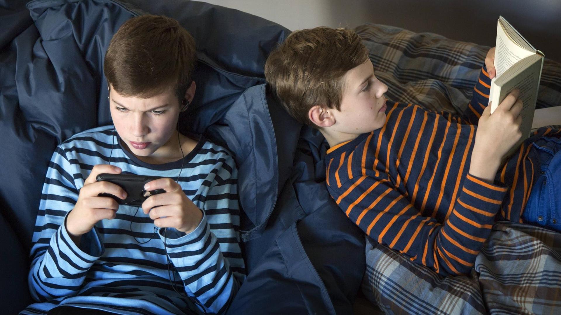 Zwei Kinder sitzen auf einer Couch - der eine spielt auf seinem Smartphone, der andere liest in einem Buch. Zwei Kinder sitzen auf einer Couch - der eine spielt auf seinem Smartphone, der andere liest in einem Buch.