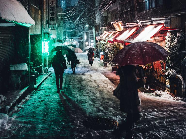 Menschen mit Regenschirmen gehen abends im Schirm durch eine kleine Straße in Tokio.