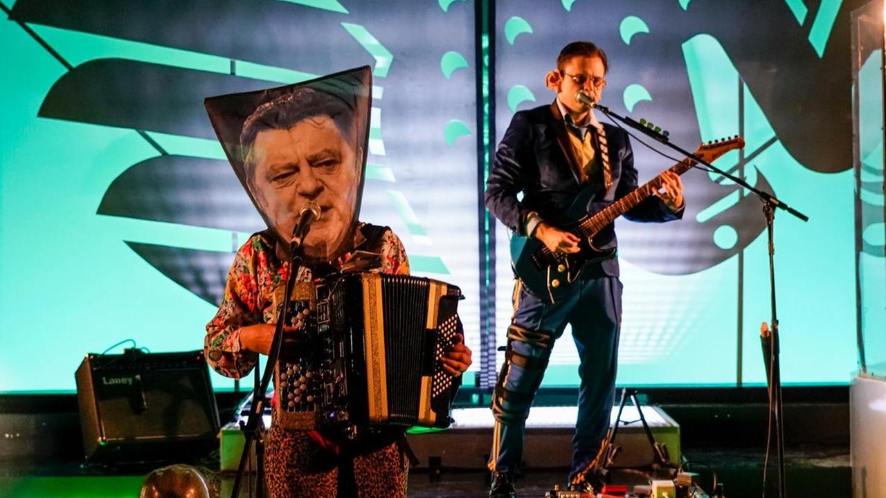 Auf einer Konzertbühne stehen zwei Musiker vor dem Hintergrund eines fallenden Bundesadlers. Einer der Musiker trägt ein Foto des früheren bayerischen Ministerpräsidenten Franz-Josef Strauß als Maske.