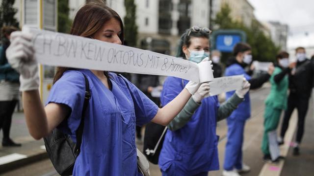 Eine medizinische Pflegekraft, in blauer Krankenhaus-Bekleding, hält während einer Demonstration ein Stück Toilettenpapier mit der Aufschrift "Ich bin kein Oktopus mit 8 Armen" in den Händen.
