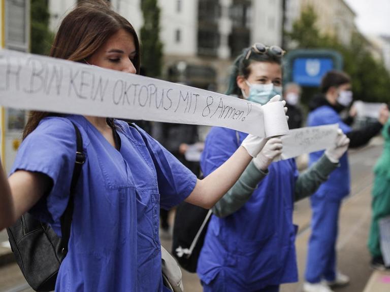Eine medizinische Pflegekraft, in blauer Krankenhaus-Bekleding, hält während einer Demonstration ein Stück Toilettenpapier mit der Aufschrift "Ich bin kein Oktopus mit 8 Armen" in den Händen.