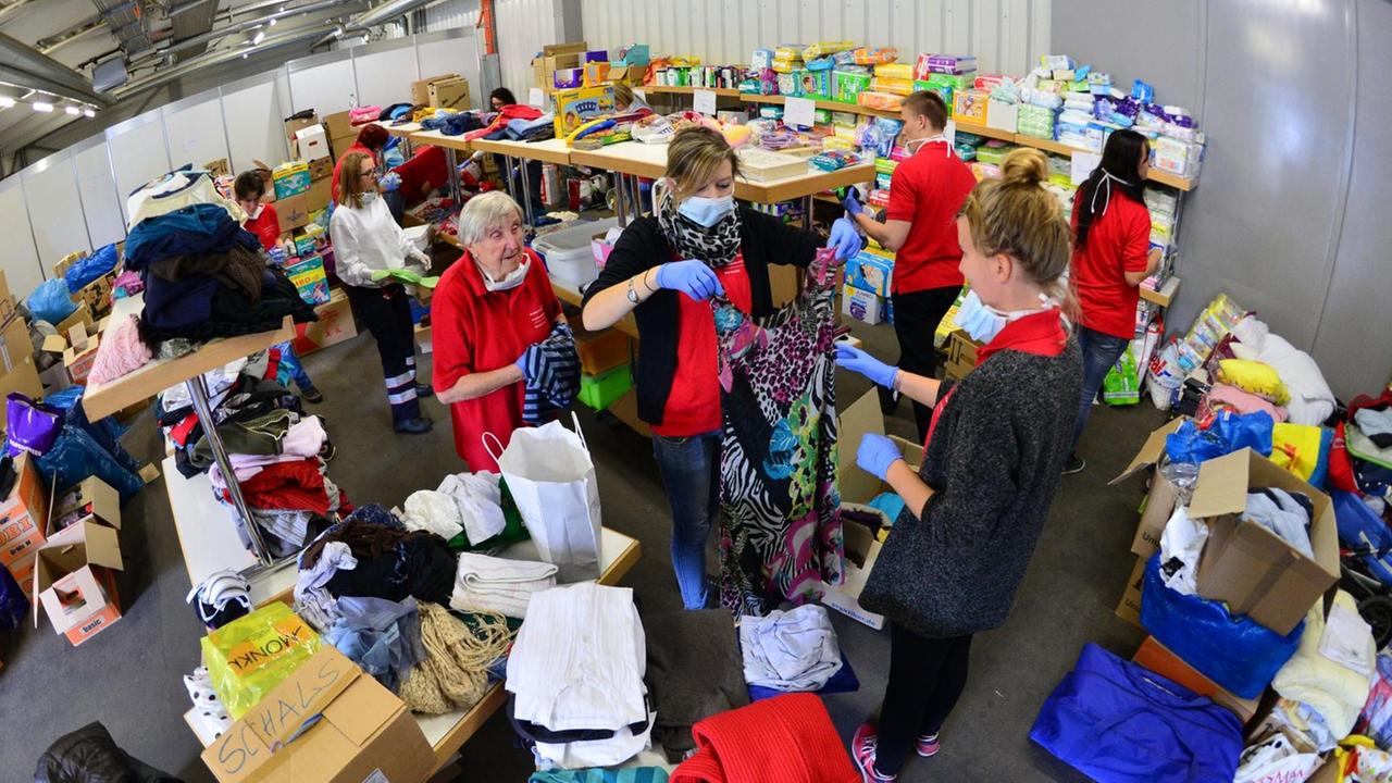 Man sieht eine Halle mit Regalen voller Kleidung und Helfer, die Kleidungsstücke sortieren. Man sieht eine Halle mit Regalen voller Kleidung und Helfer, die Kleidungsstücke sortieren.