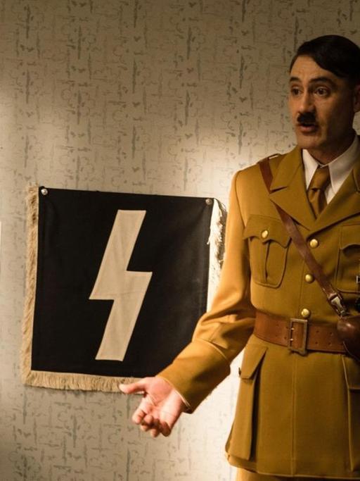 Taika Waititi steht in Nazi-Uniform und mit Hitlerbärtchen in einem Filmset.