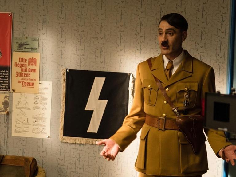 Taika Waititi steht in Nazi-Uniform und mit Hitlerbärtchen in einem Filmset.