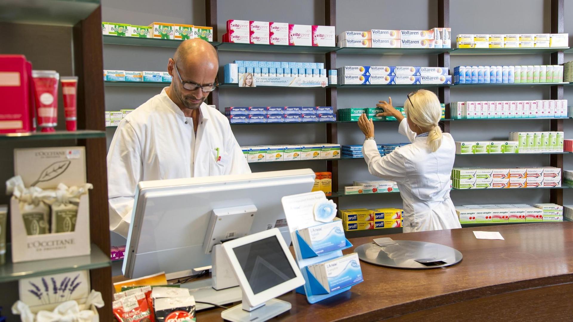 Ein Apotheker steht hinter der Ladentheke und schaut in den Computer, hinter nimmt eine Pharmazeutische Assistentin ein Medikament aus dem Regal Ein Apotheker steht hinter der Ladentheke und schaut in den Computer, hinter nimmt eine Pharmazeutische Assistentin ein Medikament aus dem Regal