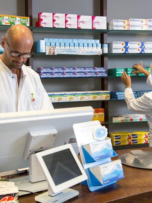 Ein Apotheker steht hinter der Ladentheke und schaut in den Computer, hinter nimmt eine Pharmazeutische Assistentin ein Medikament aus dem Regal
