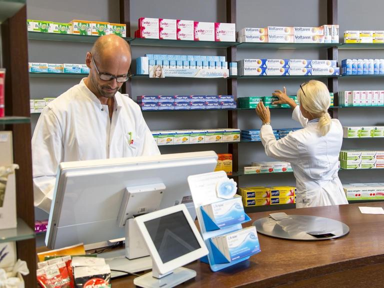 Ein Apotheker steht hinter der Ladentheke und schaut in den Computer, hinter nimmt eine Pharmazeutische Assistentin ein Medikament aus dem Regal