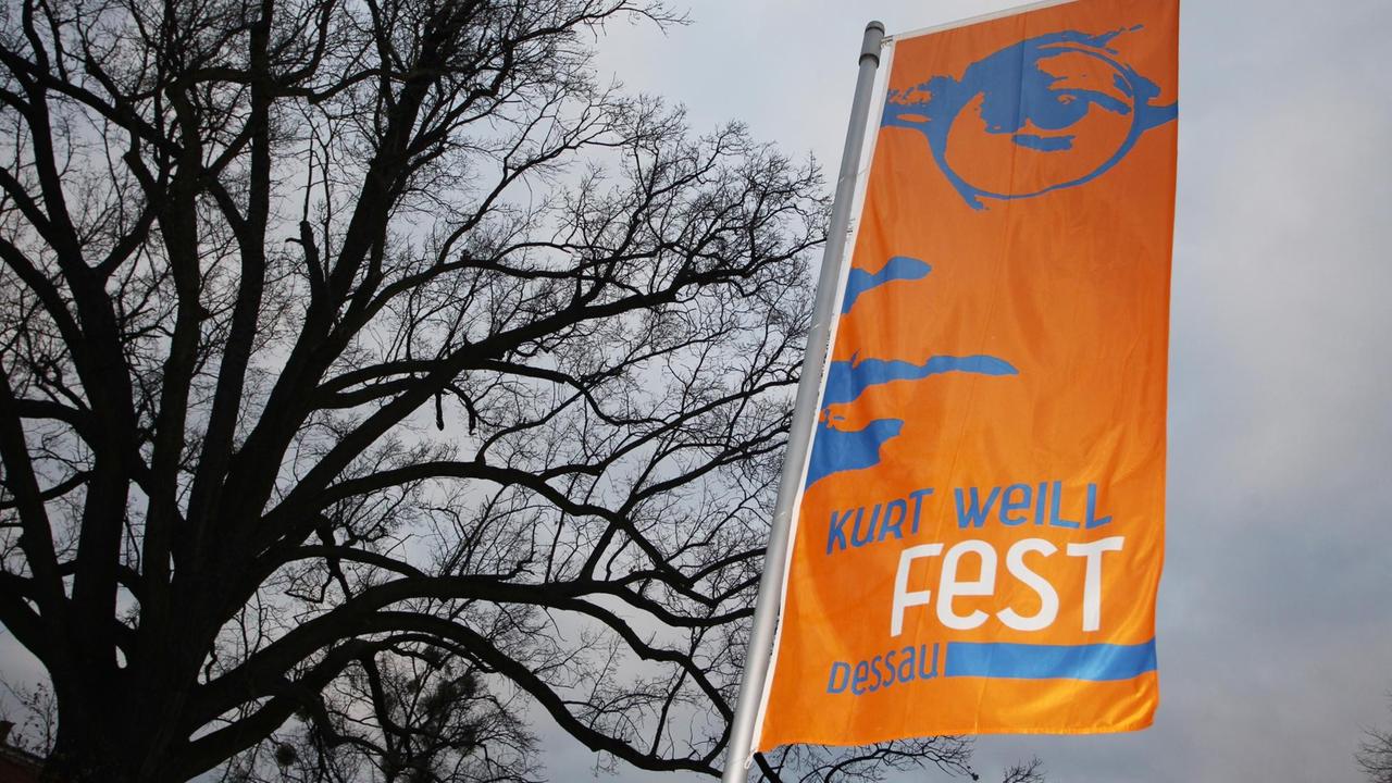 Eine orangefarbene Fane mit hellblauer Schrift weht im Wind vor einem großen, kahlen Baum. Eine orangefarbene Fane mit hellblauer Schrift weht im Wind vor einem großen, kahlen Baum.