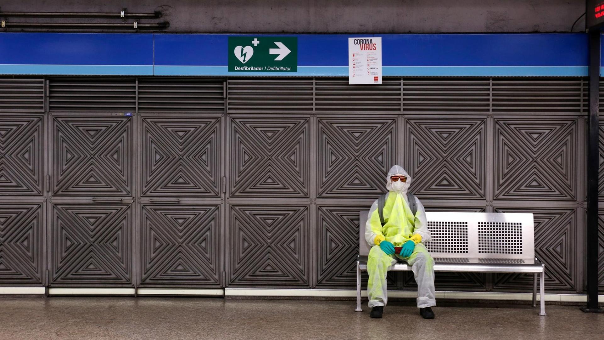 Ein Mann mit Schutzkleidung und Mundschutz sitzt in einer Metrostation in Madrid auf einer Bank. Ein Mann mit Schutzkleidung und Mundschutz sitzt in einer Metrostation in Madrid auf einer Bank.