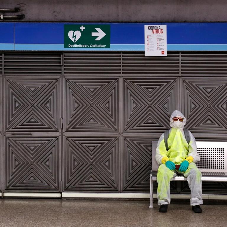 Ein Mann mit Schutzkleidung und Mundschutz sitzt in einer Metrostation in Madrid auf einer Bank. 