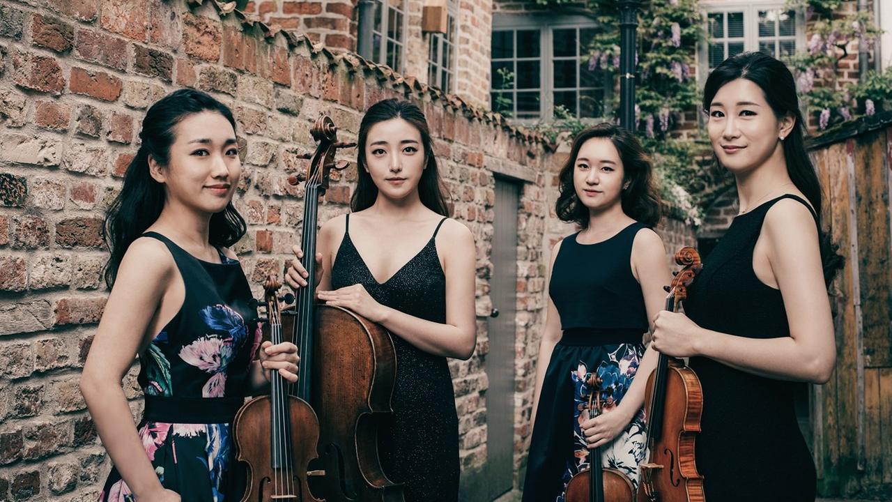 Die vier Musikerinnen stehen in blumenbedruckten schwarzen Konzertkleidern vor einer Ziegelmauer Die vier Musikerinnen stehen in blumenbedruckten schwarzen Konzertkleidern vor einer Ziegelmauer