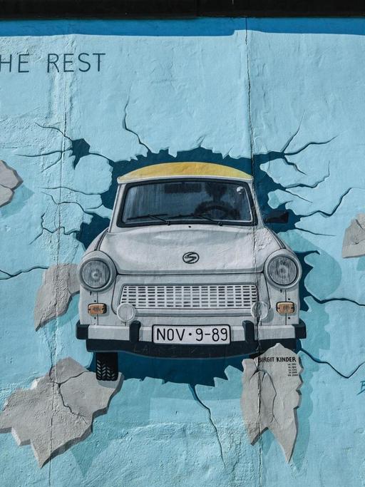 Eine der Abbildungen an der East Side Gallery in Berlin zeigt einen hellblauen Trabi, der durch eine Wand stößt.