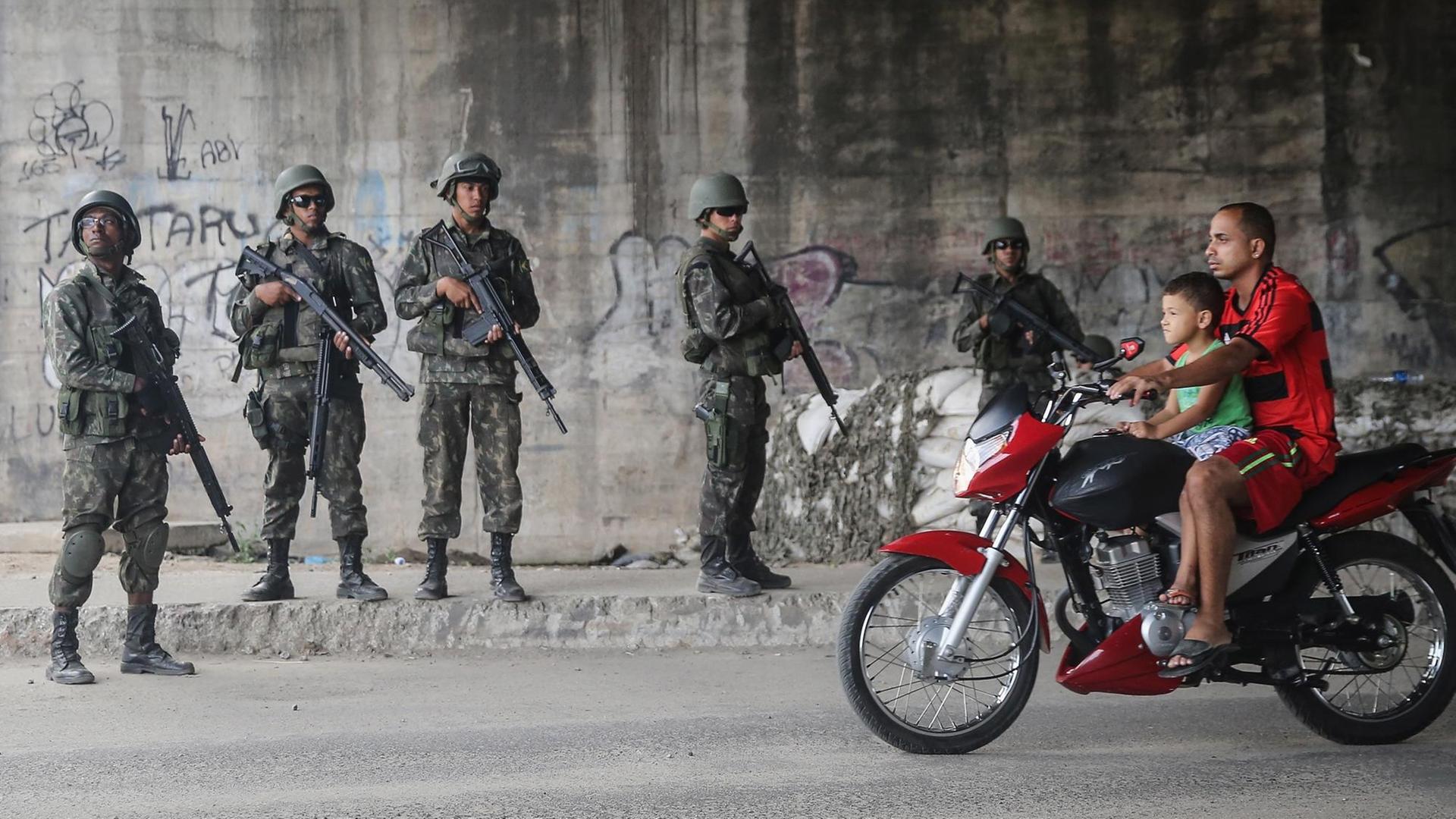 Ein Mann in der Favela Complexo da Mare in Rio de Janeiro fährt mit einem kleinen Jungen vor sich auf einem Motorrad an einer Reihe bewaffneter brasilianischer Soldaten vorbei. Ein Mann in der Favela Complexo da Mare in Rio de Janeiro fährt mit einem kleinen Jungen vor sich auf einem Motorrad an einer Reihe bewaffneter brasilianischer Soldaten vorbei.