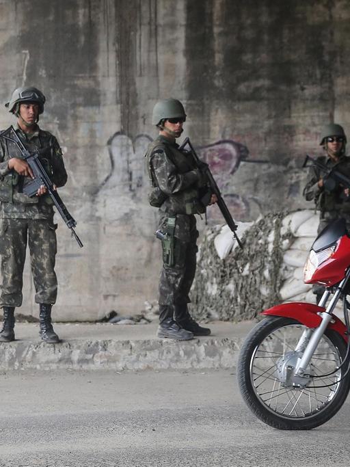 Ein Mann in der Favela Complexo da Mare in Rio de Janeiro fährt mit einem kleinen Jungen vor sich auf einem Motorrad an einer Reihe bewaffneter brasilianischer Soldaten vorbei.
