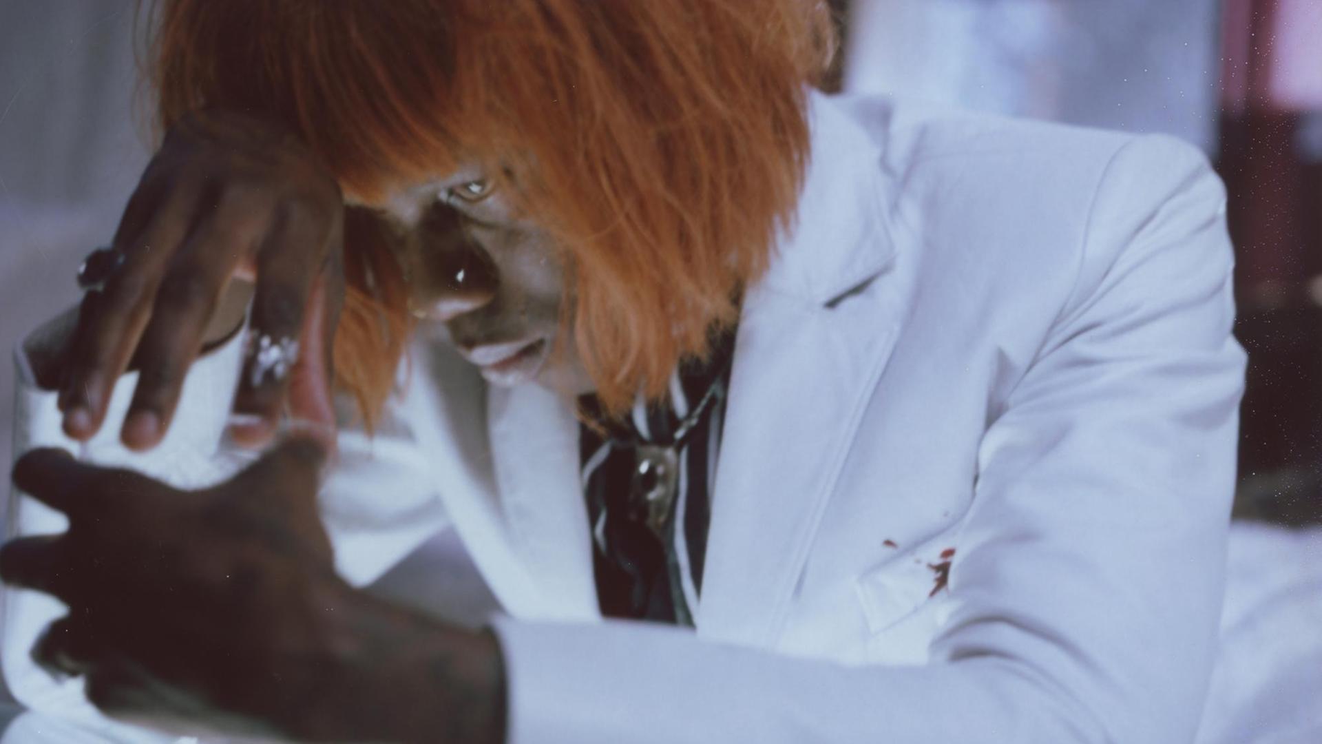 Yves Tumor posiert in einem weißen Anzug. Er trägt eine Rothaarperücke Yves Tumor posiert in einem weißen Anzug. Er trägt eine Rothaarperücke