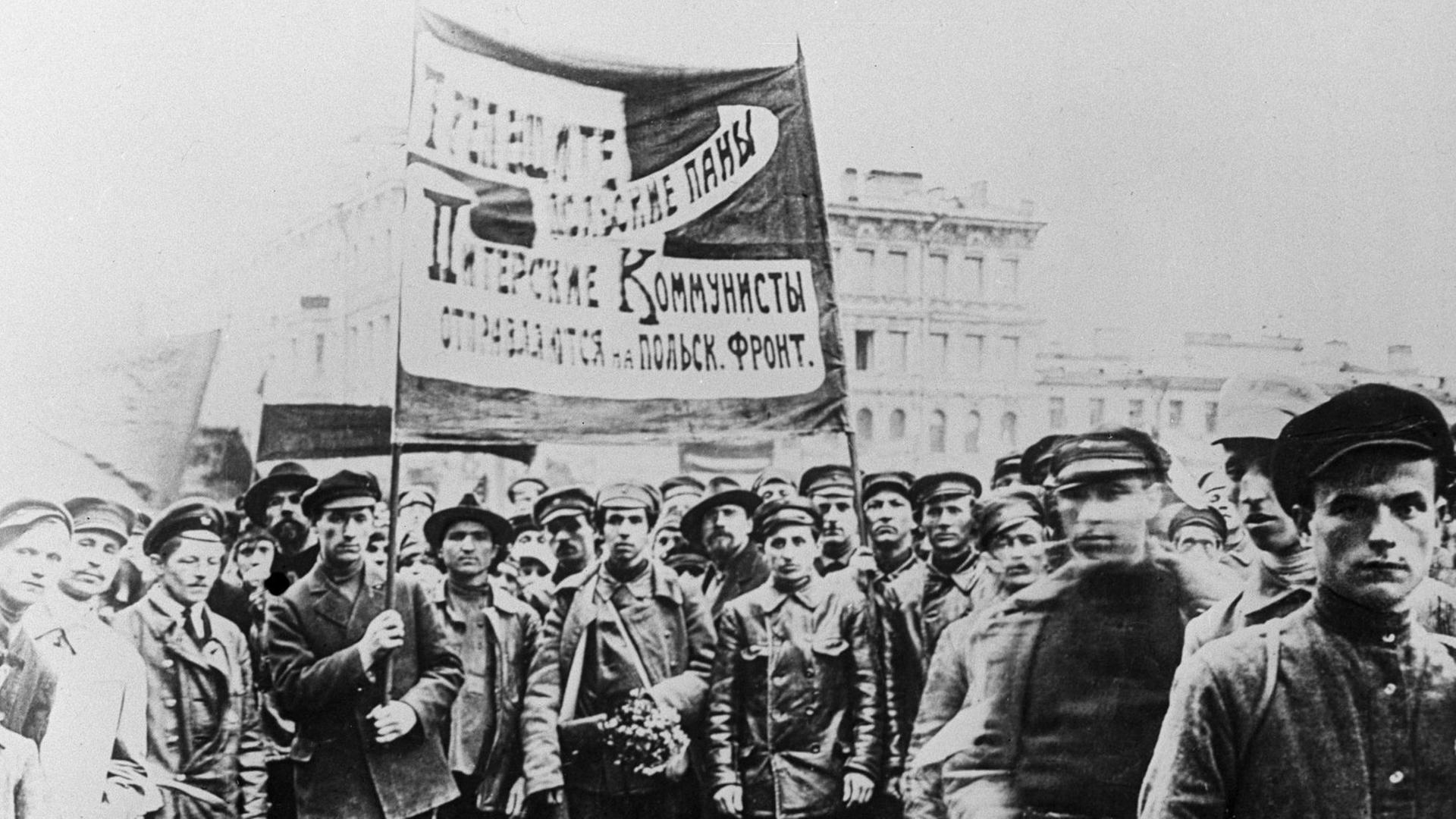 Ein Schwarzweiß-Foto zeigt etwa ein Dutzend Männer, teils in Uniform zumeist in Lederjacken und mit Schirmmützen ein großes, kyrillisch beschriebenes Banner hochhaltend Ein Schwarzweiß-Foto zeigt etwa ein Dutzend Männer, teils in Uniform zumeist in Lederjacken und mit Schirmmützen ein großes, kyrillisch beschriebenes Banner hochhaltend