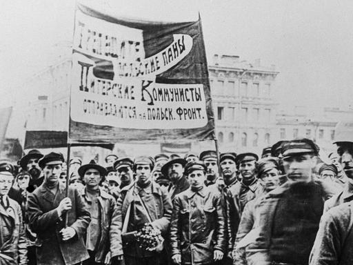 Ein Schwarzweiß-Foto zeigt etwa ein Dutzend Männer, teils in Uniform zumeist in Lederjacken und mit Schirmmützen ein großes, kyrillisch beschriebenes Banner hochhaltend