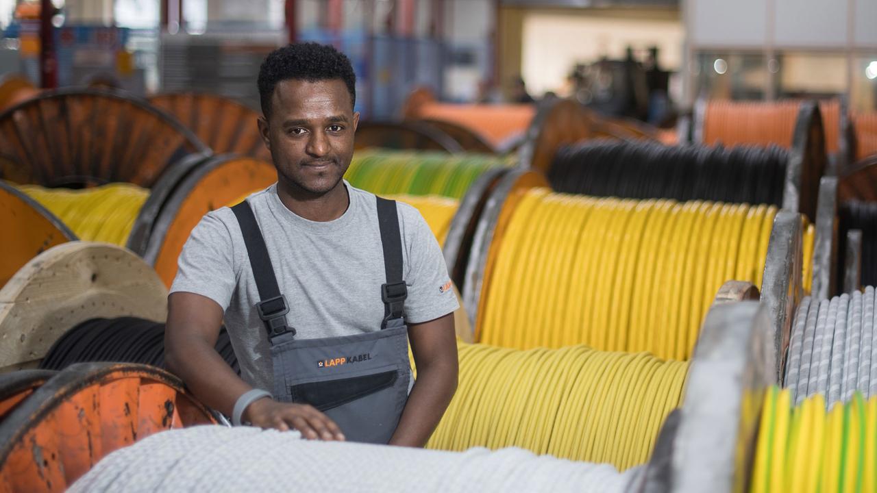 Tedros Gebru steht bei dem Kabelhersteller Lapp zwischen Kabelrollen. Nach seiner Flucht aus Eritrea hat er eine Ausbildung bei Lapp abgeschlossen und arbeitet nun in der Kabelherstellung.