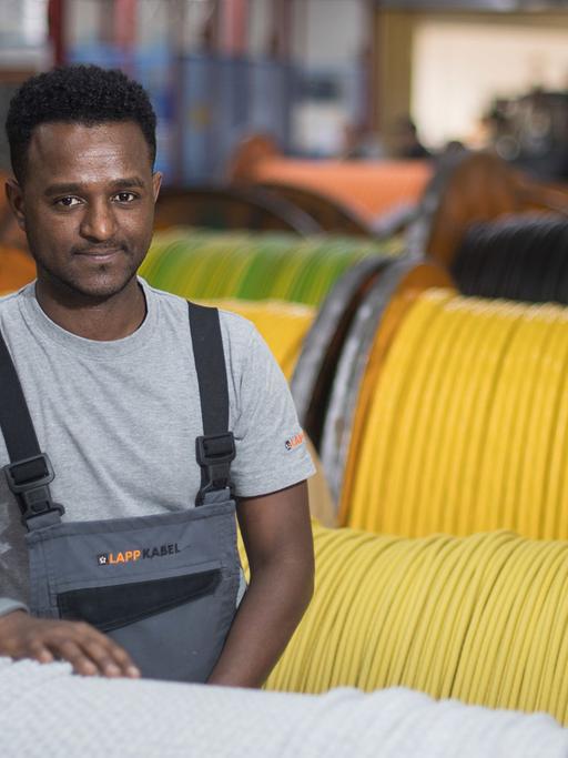 Tedros Gebru steht bei dem Kabelhersteller Lapp zwischen Kabelrollen. Nach seiner Flucht aus Eritrea hat er eine Ausbildung bei Lapp abgeschlossen und arbeitet nun in der Kabelherstellung.