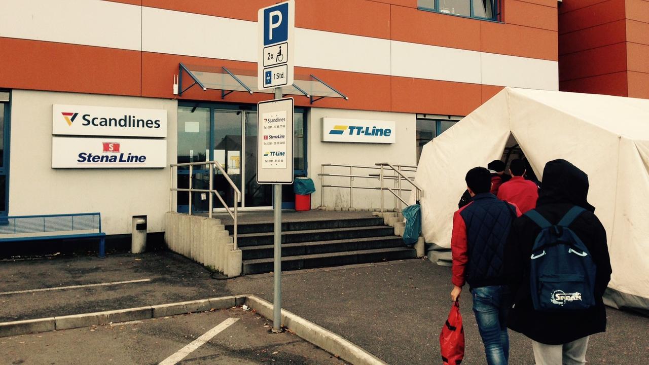 Menschen gehen in ein Zelt, das vor einem Gebäude steht, auf dem die Namen verschiedener Fährunternehmen stehen. Menschen gehen in ein Zelt, das vor einem Gebäude steht, auf dem die Namen verschiedener Fährunternehmen stehen.