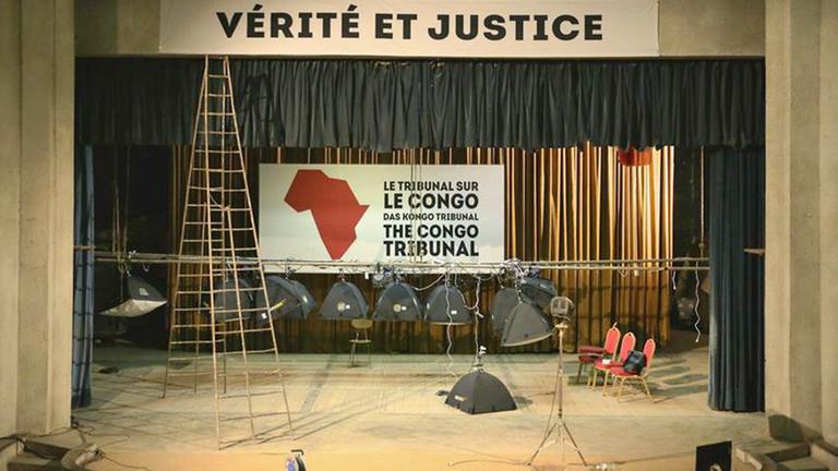 Die Ansicht einer Bühne, über der ein Plakat "Verité et Justice" schwebt, hinten eine Karte des Kongo mit Schrift "Das Kongo Tribunal" in drei Sprachen Die Ansicht einer Bühne, über der ein Plakat "Verité et Justice" schwebt, hinten eine Karte des Kongo mit Schrift "Das Kongo Tribunal" in drei Sprachen